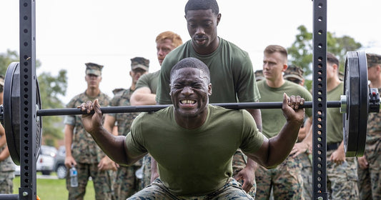 Lifter in a green T-shirt squatting a loaded free barbell in an outdoor squat rack while a spotter stands behind him