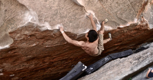 Shirtless boulderer hanging from a steep sandstone roof in the Hollow Mountain Cave, Grampians, Australia, with crash pads on the rocks below