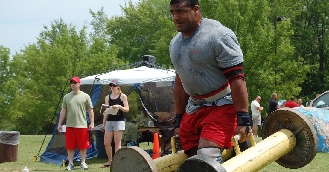 Strongman athlete mid farmer's walk, gripping the handles of two loaded carry frames on grass.