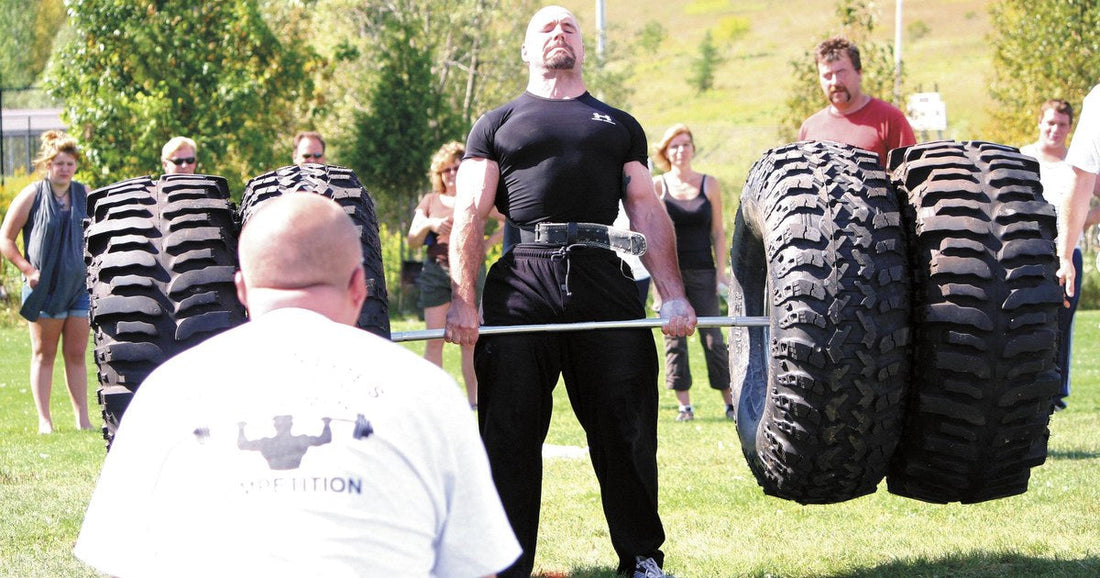 Strongman competitor in a black shirt and lifting belt locking out a tire deadlift on a grass field, with a referee kneeling in front and spectators behind