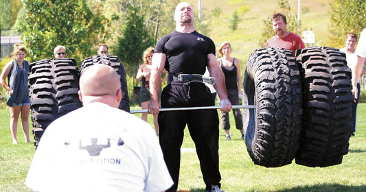 Strongman competitor in a black shirt and lifting belt locking out a tire deadlift on a grass field, with a referee kneeling in front and spectators behind